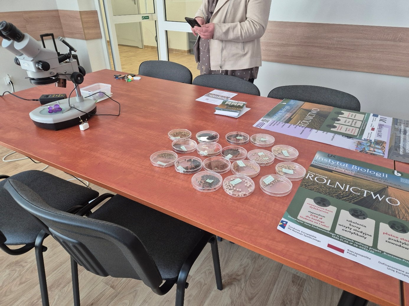 Na zdjęciu w sali lekcyjnej widoczna jest ławka szkolna, na której prezentowane są plakaty kierunku Rolnictwo oraz mikroskop z materiałem do oglądania.