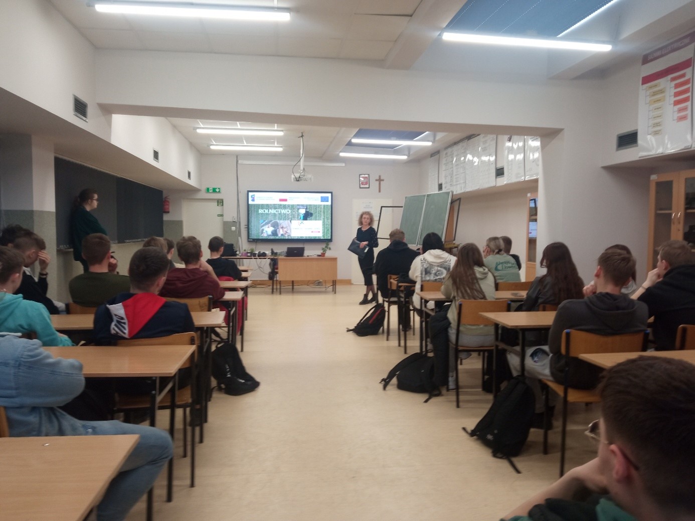Na zdjęciu w sali lekcyjnej widoczna jest młodzież szkolna oraz inne osoby uczestniczące w wykładzie. Widoczna jest także prezentacja wyświetlona na ekranie oraz kobieta prowadząca zajęcia.