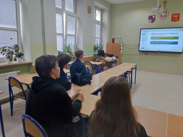 W sali lekcyjnej siedzi kilku uczniów i oglądają oni prezentację zatytułowaną Praca socjalna na UJK w Kielcach.