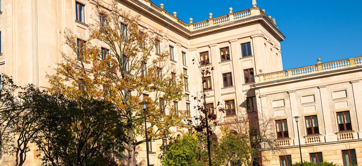 Budynek Rektoratu na tle bezchmurnego nieba, przed stoją drzewa z zielonymi liścmi