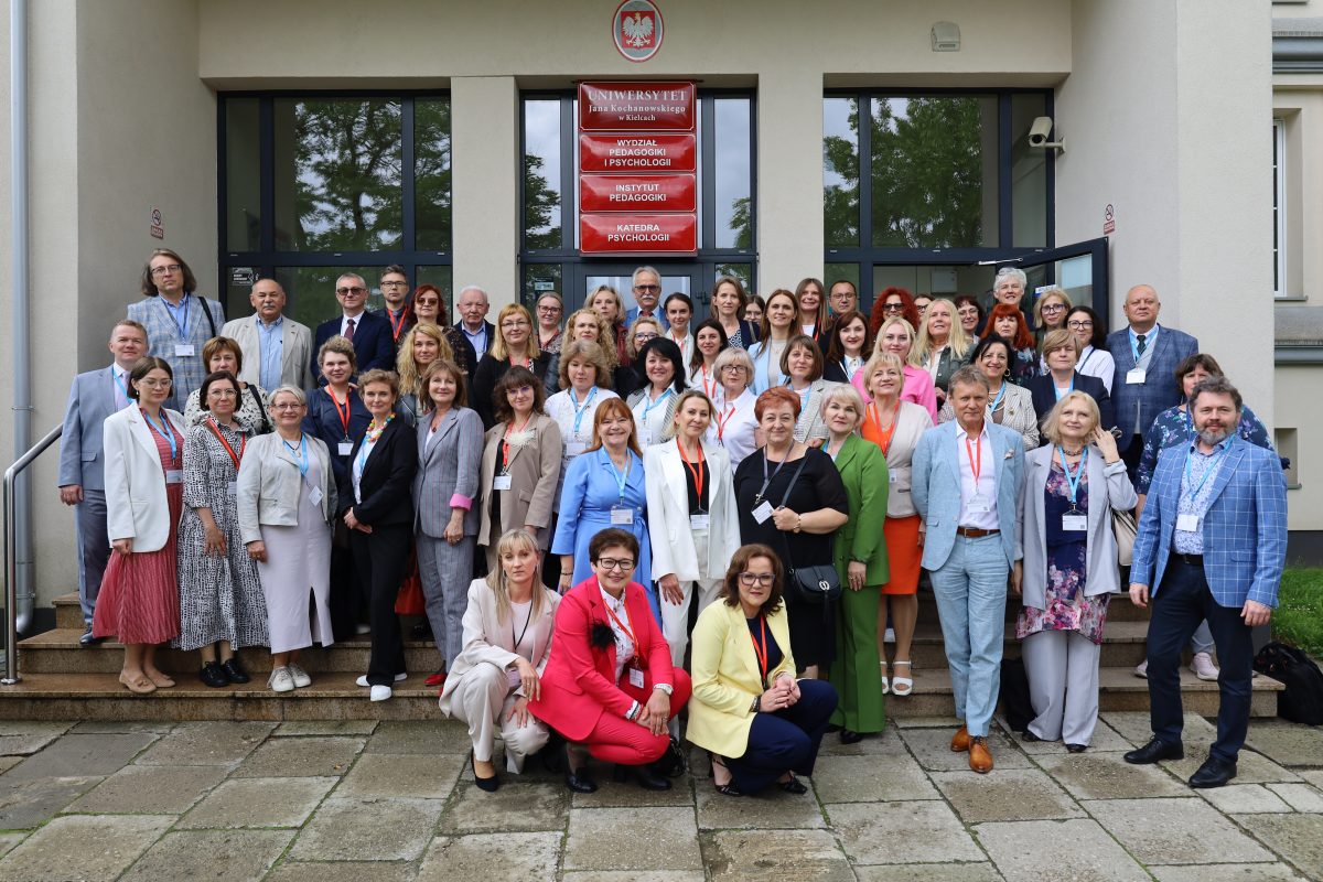 Grupowe zdjęcie dużej grupy dorosłych osób. Stoją oni i kucają na schodach przed wejściem do budynku z godłem Polski. Budynek posiada tabliczki z napisami w języku polskim, w tym "UNIWERSYTET", "WYDZIAŁ", oraz nazwy wydziałów. Mężczyźni są ubrani w garnitury, a kobiety w różnokolorowe kostiumy i sukienki. Wszyscy pozują do zdjęcia, a atmosfera wydaje się być formalna i radosna.