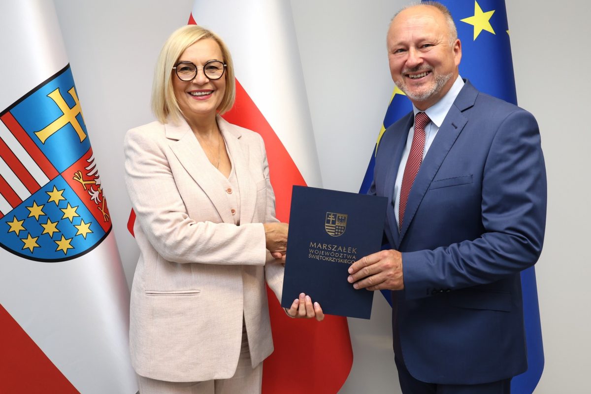 Dwie osoby - kobieta i mężczyzna stoją na tle flag Unii Europejskiej, Polski oraz flagi z herbem województwa świętokrzyskiego. Marszałek Renata Janik wręcza prof. Szymonowi Orzechowskiemu granatową teczkę z nominacją na stanowisko dyrektora.