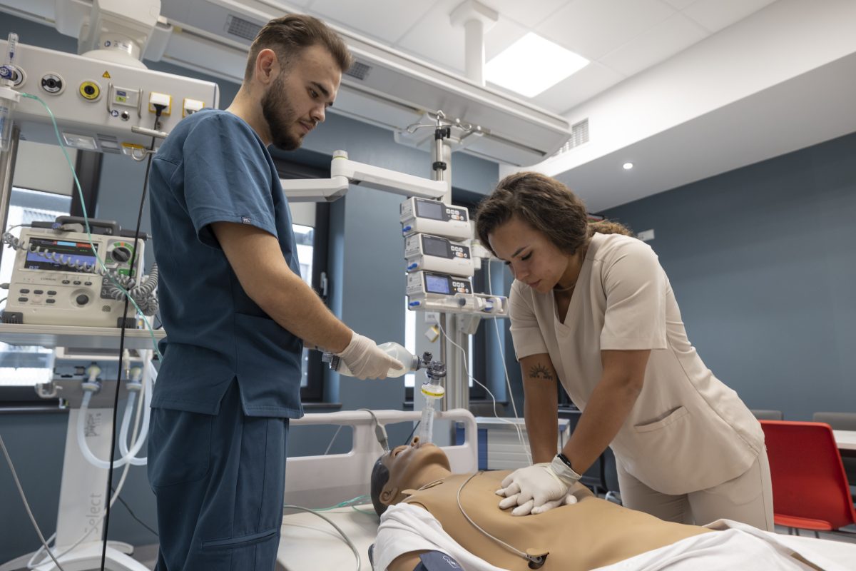 Dwoje studentów pochyla się nad fantomem do ćwiczeń. Student w niebieskim fartuchu trzyma butlę do intubacji. Studentka w białym fartuchu trzyma skrzyżowane dłonie na klatce piersiowej fantomu i wykonuje masaż serce.