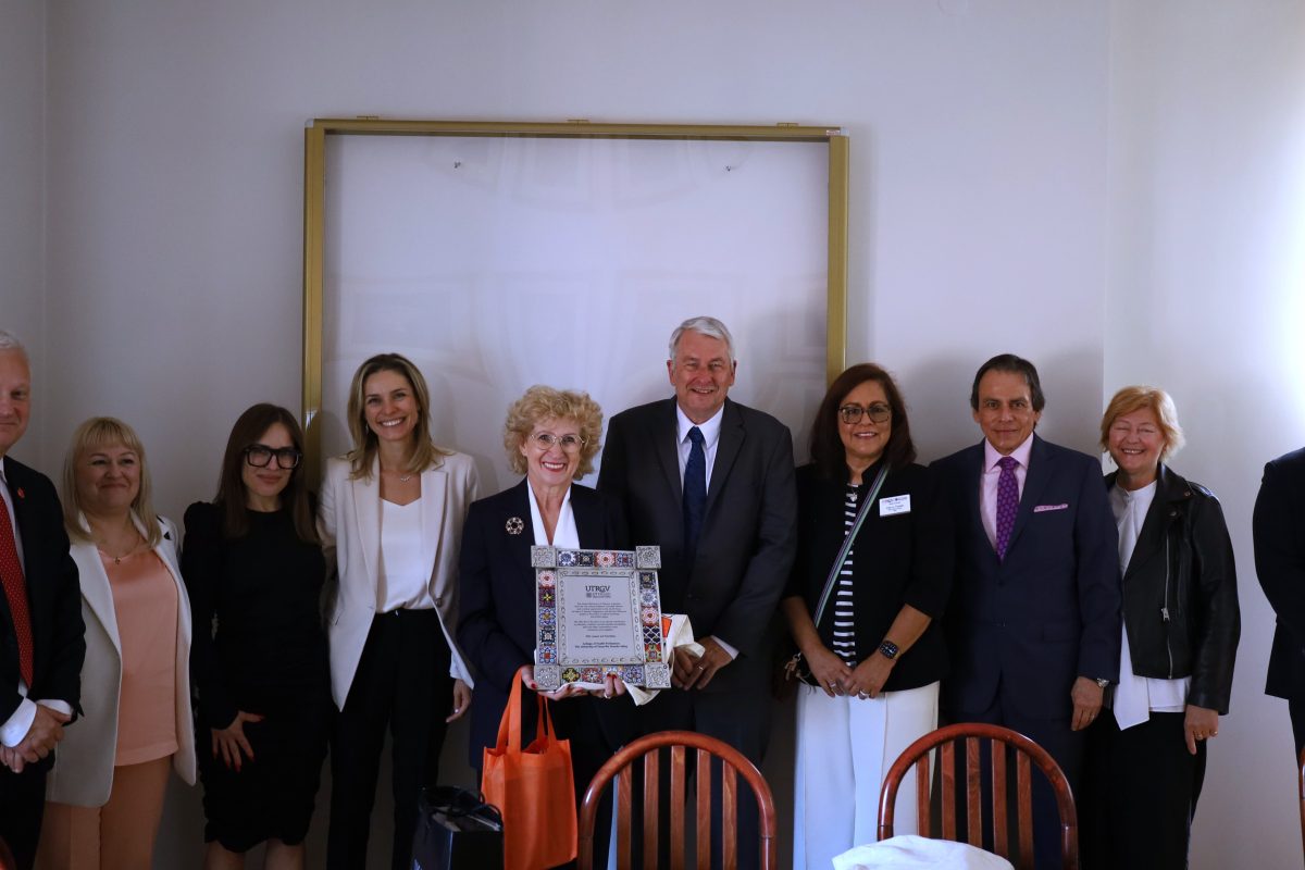 Zdjęcie grupowe dziesięciu osób w eleganckich strojach, pozujących wewnątrz jasnego pomieszczenia. W centrum kobieta o krótkich, blond włosach i wysoki, siwy mężczyzna wspólnie trzymają ozdobną, oprawioną tabliczkę. Wszyscy zebrani patrzą w kierunku aparatu, większość z nich się uśmiecha.