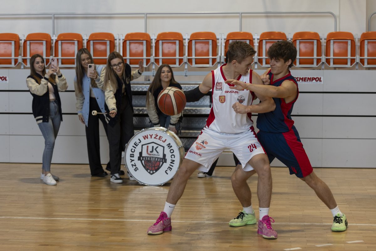 Wnętrze hali sportowej. Dwóch koszykarzy rywalizuje o piłkę. Za nimi stoją cztery dziewczyny, które dopingują zawodników.