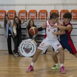 Wnętrze hali sportowej. Dwóch koszykarzy rywalizuje o piłkę. Za nimi stoją cztery dziewczyny, które dopingują zawodników.