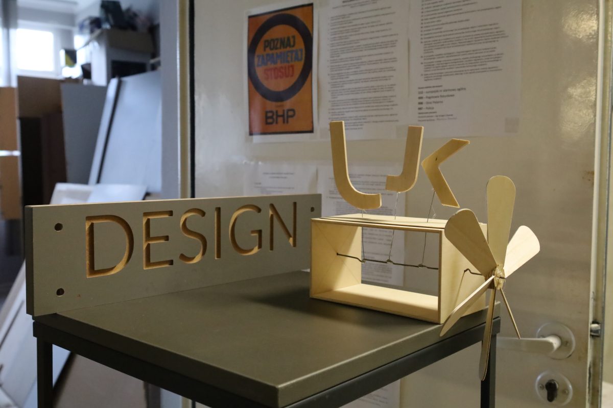 Na ciemnym stole stoją dwa drewniane obiekty. Po lewej stronie znajduje się szara tabliczka z precyzyjnie wyciętym napisem "DESIGN". Obok niej umieszczono przestrzenną kompozycję z jasnego drewna: na otwartym prostopadłościanie, na cienkich drucikach, zamocowano trzy litery i mały wiatraczek. Całość sprawia wrażenie pracowni projektowej lub warsztatu.