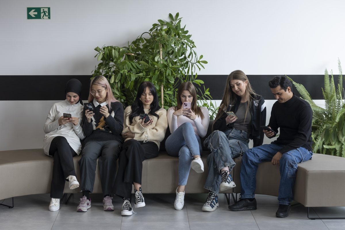 Zdjęcie przedstawia pięć osób siedzących obok siebie na ławce i wpatrujących się w ekrany swoich telefonów.