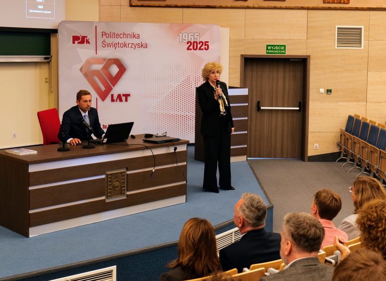 Mężczyzna siedzący przy stole i kobieta stojąca na scenie podczas prezentacji. Za nimi znajduje się baner z logo Politechniki Świętokrzyskiej i napisem "1965 2025" oraz "60 LAT". W tle widoczna jest publiczność siedząca na krzesłach.