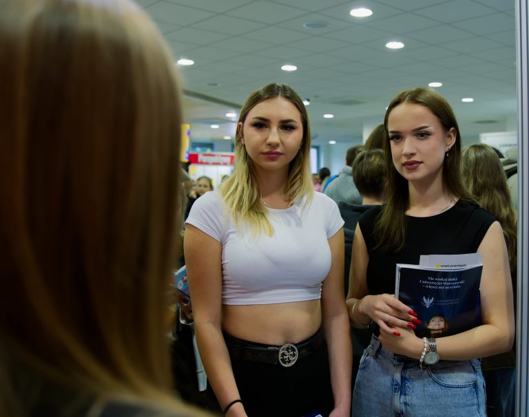 Dwie młode kobiety stoją w hali targowej, patrząc w stronę osoby rozmawiającej z nimi. Jedna ma na sobie białą koszulkę i czarne spodnie, druga czarną bluzkę i dżinsy, w rękach trzyma materiały informacyjne o uczelni.