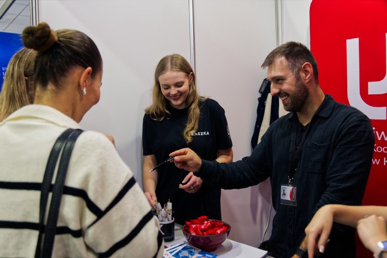 Mężczyzna z brodą i uśmiechem w ręku podaje długopis kobiecie, podczas gdy za nim uśmiecha się inna młoda kobieta, która trzyma w ręku długopis. Na stole przed nimi leży miska z czerwonymi cukierkami.