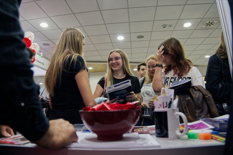 Dwie młode kobiety stoją przy stoisku informacyjnym i oglądają broszury. Jedna z nich uśmiecha się, trzymając ulotkę, a druga opiera dłoń na czole. Przed nimi, na stole, znajduje się duża miska z czerwonymi cukierkami.