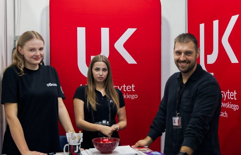Trzy osoby, dwie kobiety i jeden mężczyzna, stoją przy stoisku promocyjnym Uniwersytetu Jana Kochanowskiego w Kielcach. Za nimi widnieje duży, czerwony baner z białym logo UJK. Na stole przed nimi leży czerwona miska z cukierkami.