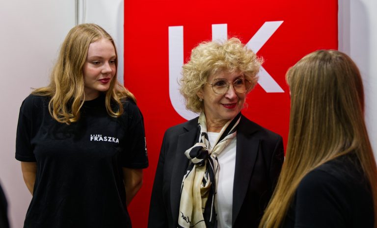 Trzy kobiety stoją i rozmawiają przy stoisku. Kobieta pośrodku z kręconymi, blond włosami i w okularach, ma na sobie czarną marynarkę i jasną apaszkę. Po jej prawej stronie stoi uśmiechnięta, młoda kobieta w czarnej koszulce, a po lewej, tyłem do kamery, druga młoda kobieta z długimi, brązowymi włosami. W tle widoczny jest czerwony baner z logo Uniwersytetu Jana Kochanowskiego.