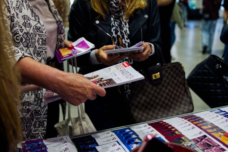 Bliskie ujęcie dłoni dwóch kobiet, które przeglądają ulotki i materiały informacyjne na stoisku. Na pierwszym planie widać materiały promocyjne z napisami w języku polskim, a w tle leży torebka w szachownicę.