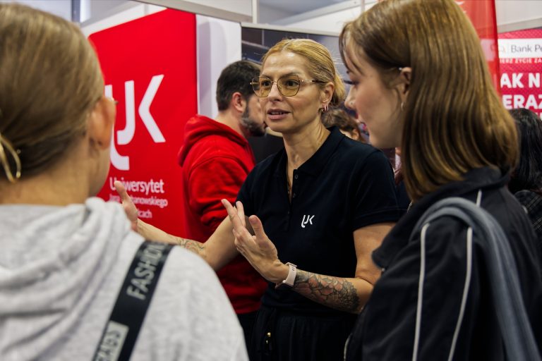 Kobieta z tatuażami na rękach, w okularach i czarnej koszulce z logo UJK, rozmawia z dwiema młodymi osobami na stoisku uniwersytetu. Używa gestykulacji, próbując przekazać jakieś informacje. W tle widoczny jest mężczyzna w czerwonej bluzie i fragment baneru reklamowego. Analiza