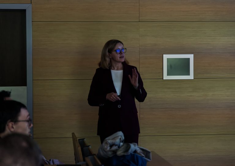 Kobieta w granatowej marynarce i białej koszulce, w okularach, stoi na tle drewnianej ściany i przemawia, gestykulując lewą ręką.