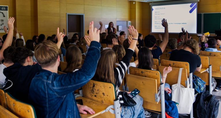 Szerokie ujęcie z tyłu sali wykładowej, gdzie duża grupa młodych ludzi siedzi na drewnianych ławkach. Wiele osób ma podniesione ręce. Z przodu sali, na ekranie, widać slajd z napisem „AGENDA”. W tle widoczna jest kobieta, prawdopodobnie wykładowca.