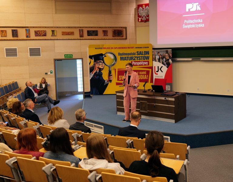 Kobieta stojąca na scenie w różowym garniturze przemawia do publiczności. Na stole przed nią leży laptop. Za nią widać banery z napisami "PSK Politechnika Świętokrzyska" i "Ogólnopolski Salon Maturzystów". Na ekranie wyświetla się prezentacja z logo Politechniki Świętokrzyskiej.