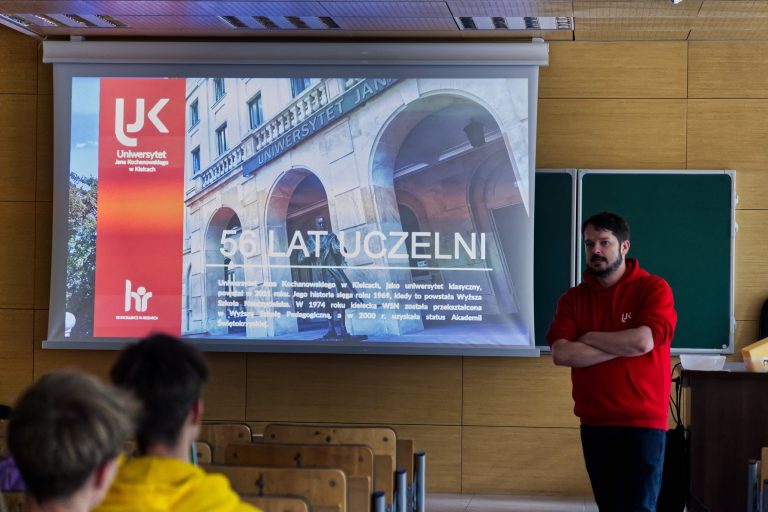 Mężczyzna z brodą w czerwonej bluzie stoi na scenie, ze skrzyżowanymi rękami. Obok niego, na ekranie, wyświetla się prezentacja z napisem "UNIWERSYTET JANA KOCHANOWSKIEGO" i "56 LAT UCZELNI". Na slajdzie widać również zdjęcie budynku z arkadami oraz logo uczelni.