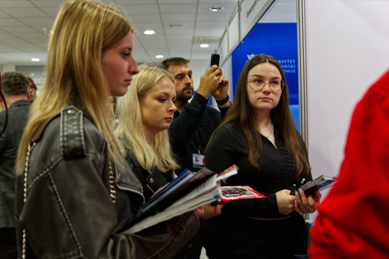 Trzy młode osoby stoją i przeglądają materiały promocyjne na stoisku. Dwie z nich trzymają w rękach broszury i ulotki. Za nimi w tle widać brodatego mężczyznę, który trzyma telefon, prawdopodobnie robiąc zdjęcie.