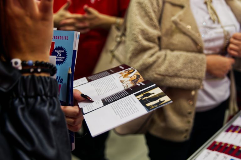 Kobiety trzymają w rękach broszury i ulotki, przeglądając je podczas targów edukacyjnych. Jedna z nich ma pomalowane na czarno paznokcie. W tle widać inne osoby, które również oglądają materiały.