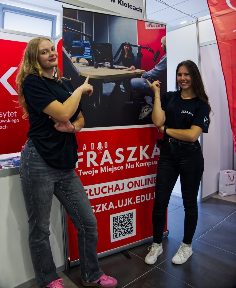 Dwie młode kobiety, ubrane w czarne koszulki z napisem "FRASZKA", pozują przy banerze promocyjnym radiostacji. Obie wskazują kciukami w stronę baneru, a jedna z nich ma na sobie jasne spodnie, a druga ciemne. Baner przedstawia zdjęcie z wnętrza studia radiowego i zachęca do słuchania online.