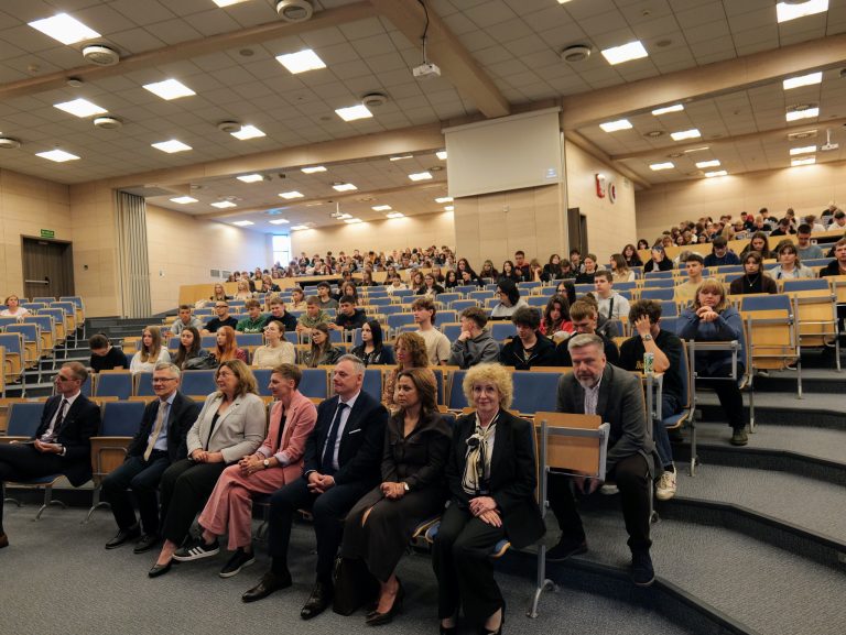 Publiczność siedząca w dużym audytorium. W pierwszych rzędach siedzi grupa dorosłych, a za nimi, w dalszej części sali, widać wielu młodych ludzi.