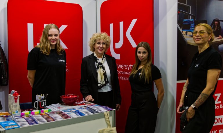 Cztery kobiety stoją przy stoisku promocyjnym. Na czerwonym tle za nimi widnieje logo Uniwersytetu Jana Kochanowskiego w Kielcach. Na stole przed nimi leżą ulotki i materiały informacyjne.