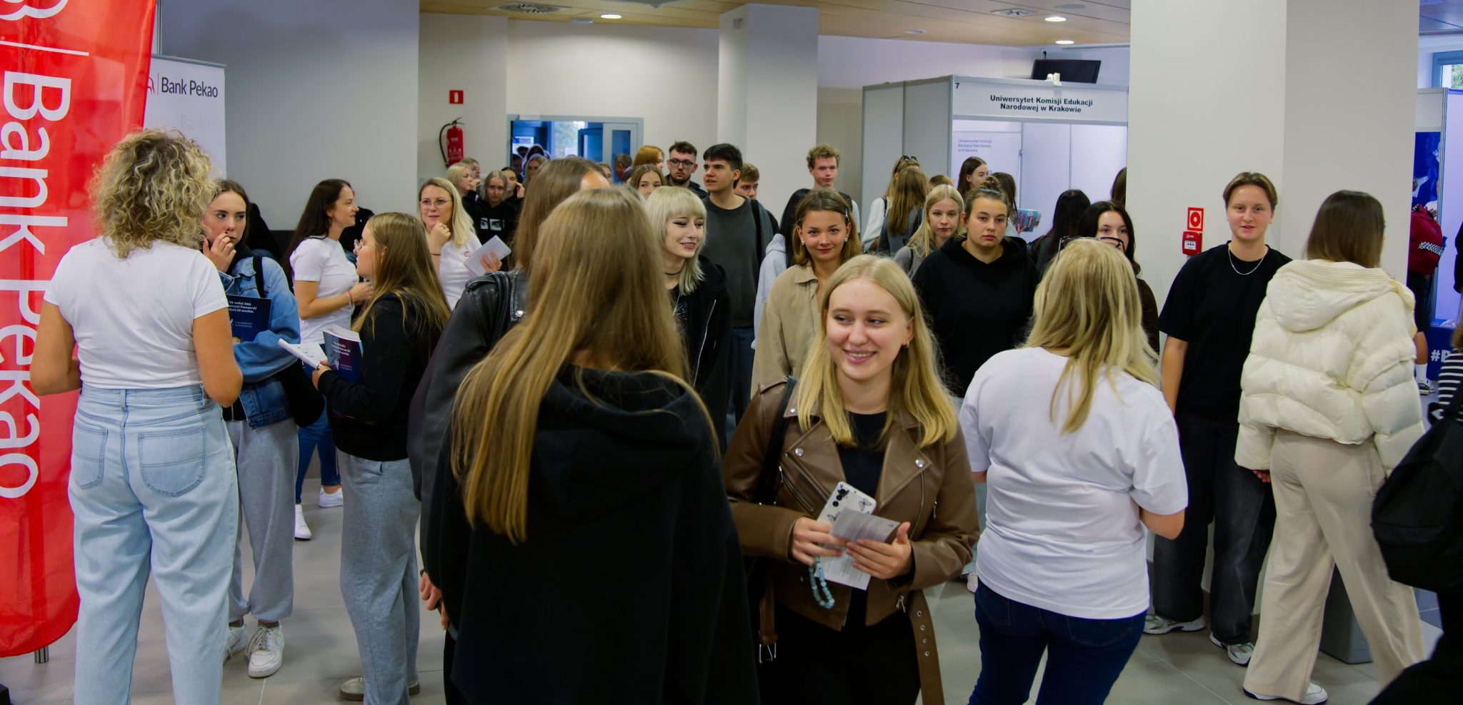 Duża grupa młodych ludzi, prawdopodobnie studentów, stoi na korytarzu podczas wydarzenia, rozmawiając ze sobą i oglądając stoiska. W tle widoczne są banery promocyjne, w tym jeden z logo Banku Pekao.
