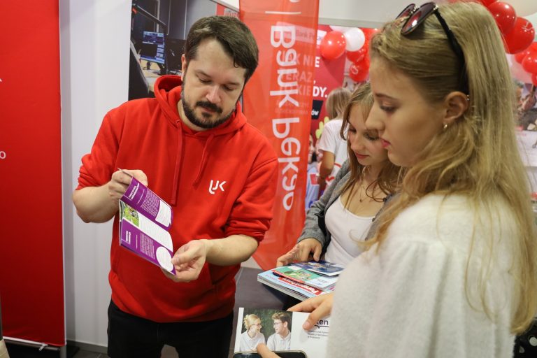 Mężczyzna w czerwonej bluzie z kapturem, stojący za stoiskiem promocyjnym, pokazuje ulotkę dwóm młodym kobietom, które przyglądają się materiałom informacyjnym. W tle widoczne są czerwone i białe balony oraz baner z napisem „Bank Pekao”.