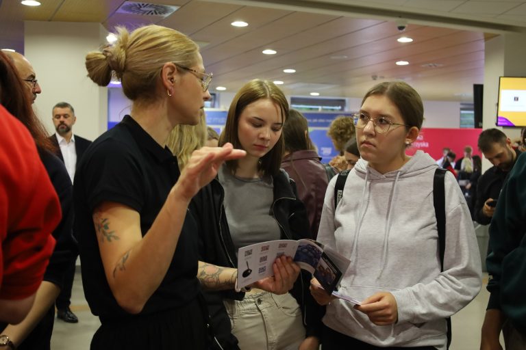 „Kobieta w okularach i czarnej koszulce rozmawia z dwiema młodymi osobami w przestrzeni wystawowej. Jedna z nich trzyma otwartą ulotkę, druga ma na sobie jasną bluzę i plecak. W tle widać innych uczestników targów.”