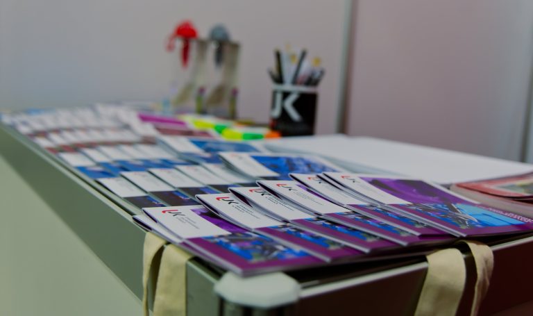 Zblizenie na stół z poukładanymi w rzędach ulotkami i broszurami. Na pierwszym planie widać broszury z logo Uniwersytetu Jana Kochanowskiego (UJK). W tle znajdują się markery i pojemnik na długopisy.