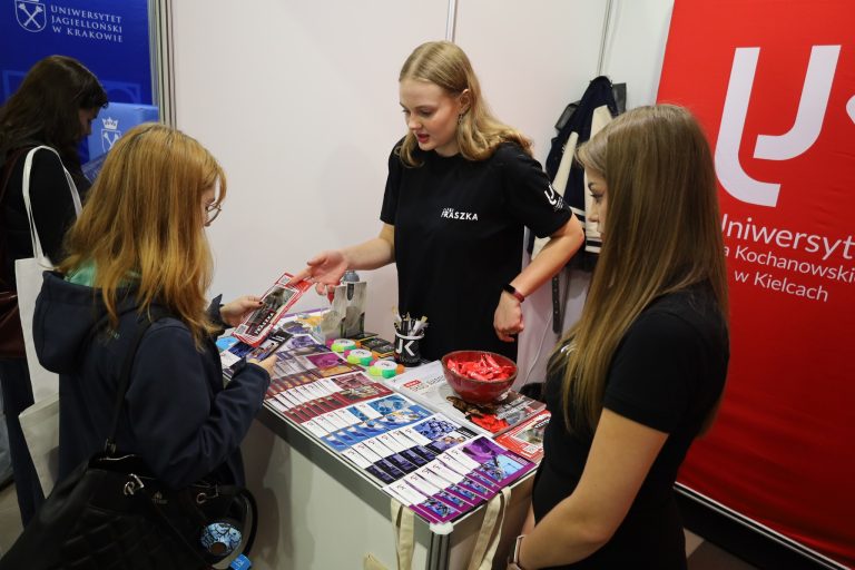 Dwie młode kobiety stoją przy stoisku informacyjnym i oglądają broszury. Jedna z nich uśmiecha się, trzymając ulotkę, a druga opiera dłoń na czole. Przed nimi, na stole, znajduje się duża miska z czerwonymi cukierkami.