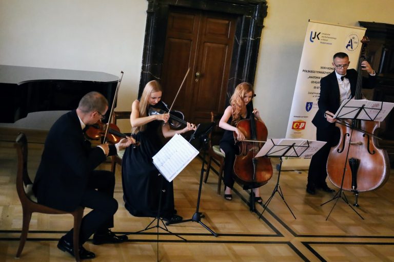 Na zdjęciu widać kwartet smyczkowy, składający się z dwóch mężczyzn i dwóch kobiet. Mężczyzna siedzący po lewej stronie gra na skrzypcach. Obok niego, w centrum, siedzą dwie kobiety. Jedna gra na skrzypcach, a druga na wiolonczeli. Po prawej stronie zdjęcia stoi mężczyzna grający na kontrabasie. Wszyscy są ubrani w czarne, eleganckie stroje. Muzycy grają w jasnym pomieszczeniu. W tle widoczny jest fortepian, a za kontrabasistą znajduje się baner reklamowy.