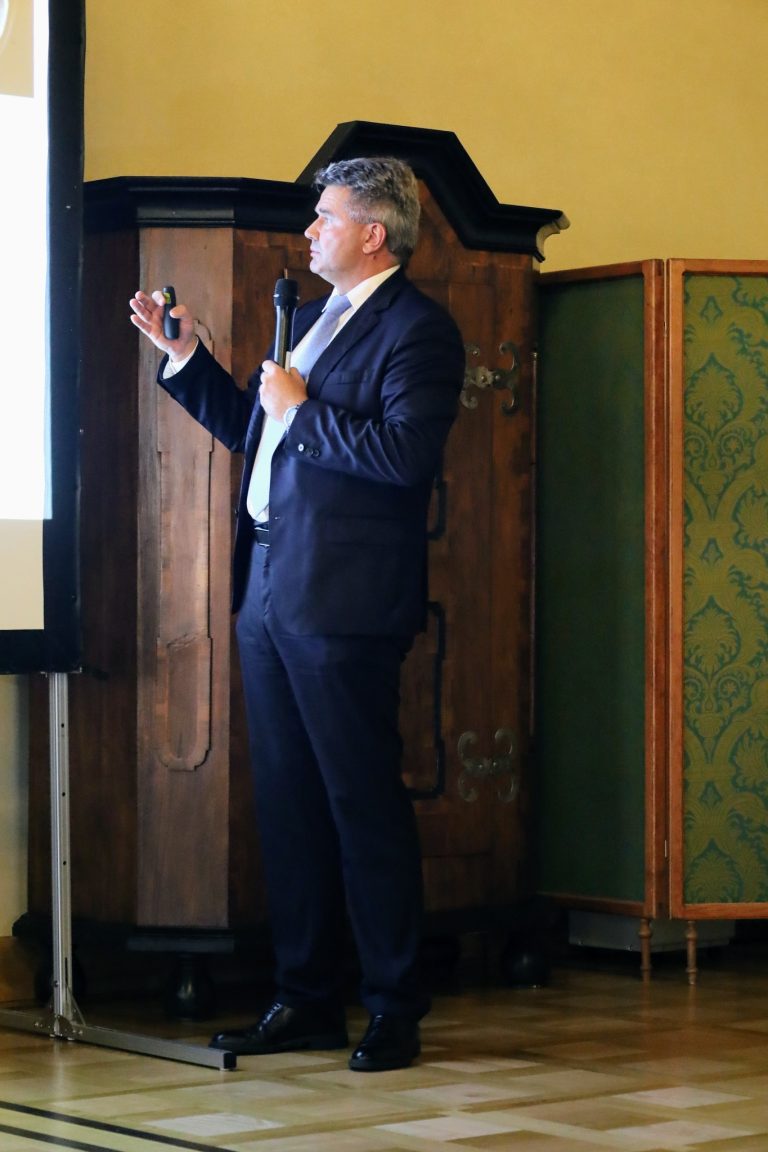 Na zdjęciu jest mężczyzna, który ma na sobie ciemny garnitur i jasny krawat. Stoi obok ekranu projekcyjnego, na którym wyświetlana jest prezentacja. W lewej ręce trzyma pilot do slajdów, a w prawej mikrofon. Mężczyzna jest zwrócony w lewą stronę, patrząc na ekran. W tle widoczna jest zabytkowa, drewniana szafa oraz parawan z wzorzystego materiału.