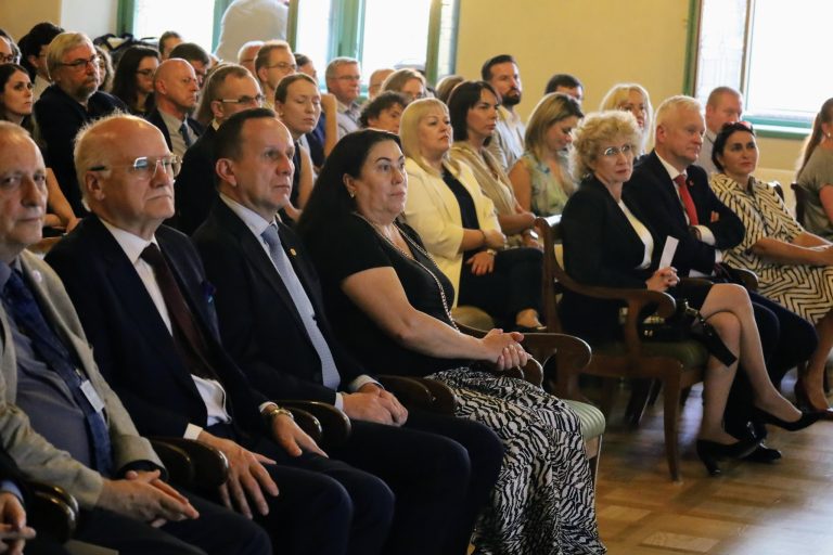 Na zdjęciu widoczna jest publiczność siedząca w rzędach krzeseł i słuchająca prelekcji. W pierwszym rzędzie, od lewej, siedzi kilku mężczyzn w garniturach, a obok nich kobieta w czarnej bluzce i spódnicy z wzorem. Jej ręce są złożone na kolanach. Dalej w rzędzie siedzi starsza kobieta w czarnym żakiecie, trzymająca w dłoni biały papier. W tle widać kolejnych uczestników, w większości w eleganckich strojach. Wszyscy siedzą, patrząc w jednym kierunku, prawdopodobnie w stronę sceny, która jest poza kadrem.