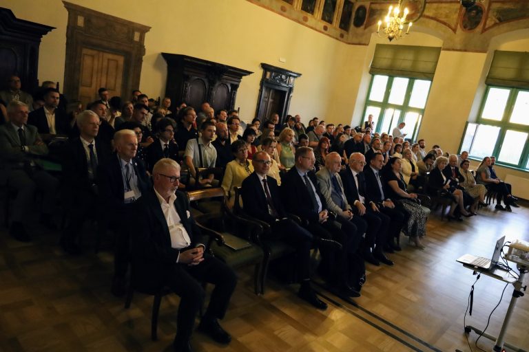 Na zdjęciu widoczna jest duża grupa ludzi, siedzących w rzędach krzeseł w jasnej, zabytkowej sali. Publiczność jest zwrócona w stronę, która jest poza kadrem. W pierwszym rzędzie po lewej stronie siedzi mężczyzna w czarnym garniturze i białej koszuli, spoglądający w stronę obiektywu, podczas gdy inni uczestnicy patrzą w tym samym kierunku. Na podłodze sali widać drewniane panele. W tle, na ścianach, wiszą obrazy i widać zabytkowe, drewniane meble. Duże okna wpuszczają do pomieszczenia dużo światła.