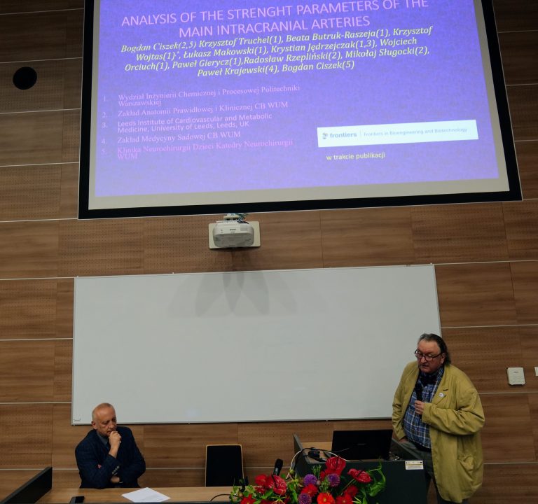 a zdjęciu widać salę konferencyjną, a na pierwszym planie są dwaj mężczyźni. Po lewej stronie siedzi mężczyzna w granatowej marynarce, podpierający brodę ręką. Po prawej, stojąc, znajduje się mężczyzna z długimi włosami i brodą, ubrany w jasną marynarkę, trzymający mikrofon w ręce. W tle, na ścianie, wyświetlana jest prezentacja z nagłówkiem: "ANALYSIS OF THE STRENGTH PARAMETERS OF THE MAIN INTRACRANIAL ARTERIES". Pod nagłówkiem znajduje się lista nazwisk i afiliacji. Na stole, przed siedzącym mężczyzną, widać bukiet kwiatów i laptopa.
