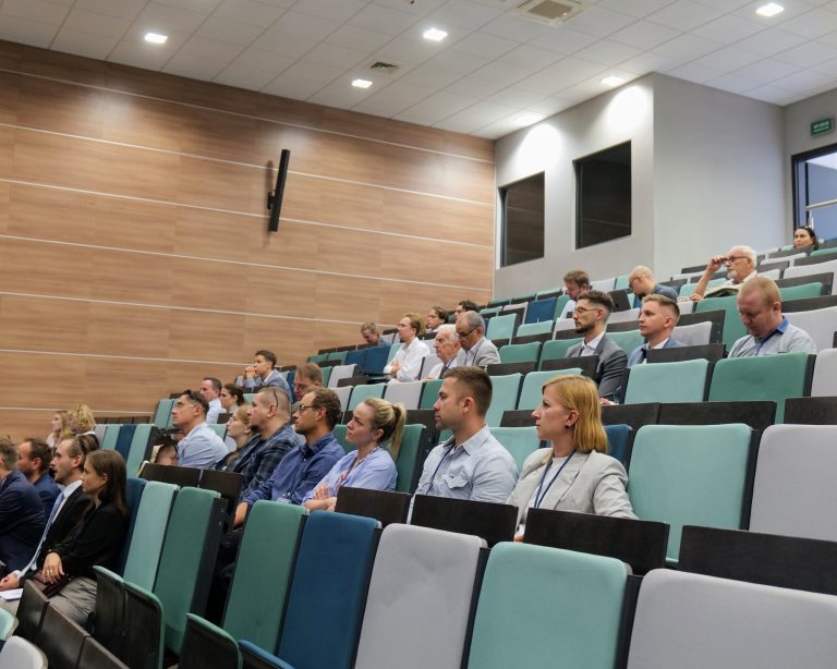 Na zdjęciu widoczna jest duża grupa osób, siedzących w rzędach krzeseł w sali wykładowej lub audytoryjnej. Krzesła są w kilku odcieniach zielonego i niebieskiego. Większość uczestników jest zwrócona w jednym kierunku, prawdopodobnie słuchając prelegenta, który jest poza kadrem. W tle widać ściany pokryte drewnianymi panelami i włączone oświetlenie.