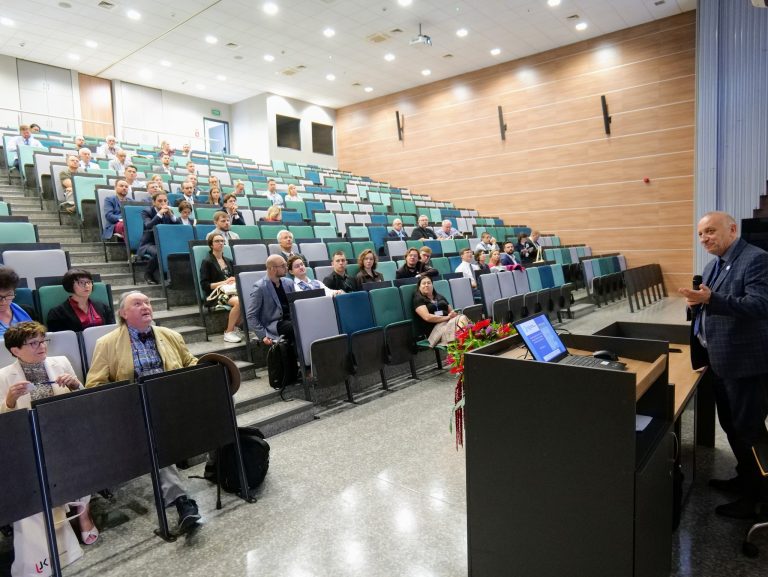 Na zdjęciu widoczna jest duża sala wykładowa, wypełniona rzędami krzeseł, na których siedzi publiczność. Po prawej stronie kadru, przy pulpicie, stoi mężczyzna w granatowej marynarce w kratę, uśmiecha się i gestykuluje rękoma w stronę publiczności. Publiczność, w większości, patrzy w kierunku prelegenta. Sala ma stopniowane rzędy, a na pulpicie widoczny jest laptop. W tle, na ścianie, wiszą dekoracyjne panele.