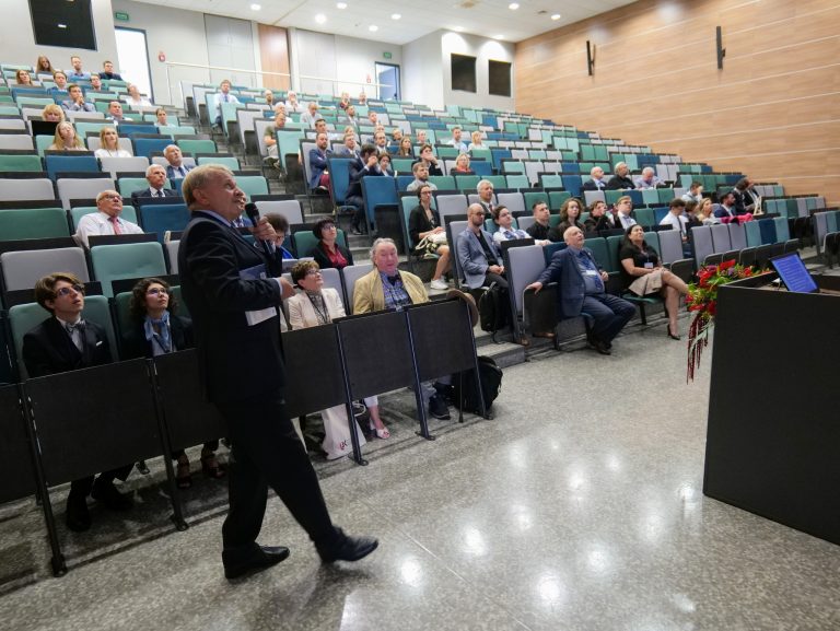 Na zdjęciu widoczna jest duża sala wykładowa, wypełniona publicznością siedzącą w rzędach. Na pierwszym planie, po lewej stronie, stoi mężczyzna w granatowym garniturze, trzymający w ręce mikrofon. Spogląda w stronę publiczności, gestykulując w trakcie przemówienia. Na środku sali, na niższych rzędach, widać słuchaczy. Z kolei w rzędach wyżej, na podwyższeniu, siedzą pozostali uczestnicy. Sala ma podłogę pokrytą szarymi panelami, a na ścianach widać drewniane okładziny.
