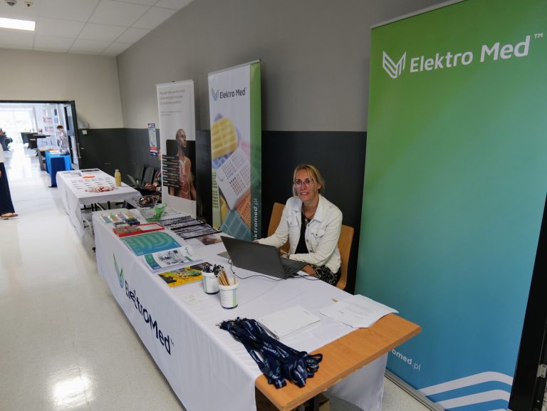 Na zdjęciu widoczna jest kobieta siedząca za stoiskiem wystawienniczym firmy Elektro Med. Kobieta ma na sobie białą bluzę i okrągłe okulary, uśmiecha się do obiektywu. Przed nią, na stole nakrytym białym obrusem z logo firmy, leży laptop, długopisy, notatniki, broszury i materiały promocyjne. Po obu stronach stoiska stoją pionowe banery reklamowe z logo firmy. W tle widać fragment korytarza oraz inne, częściowo ukryte stoiska.