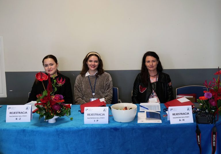 Na zdjęciu widoczne są trzy kobiety siedzące za długim stołem, który służy jako punkt rejestracyjny. Stół jest nakryty niebieskim obrusem. Przed każdą z kobiet leżą tabliczki z napisem "REJESTRACJA" i zakresami liter dla nazwisk: "R-Z", "I-P" i "A-H". Kobiety uśmiechają się. Po lewej i prawej stronie stołu stoją bukiety kwiatów, a na środku widoczna jest biała miska z jedzeniem. Kobiety są ubrane swobodnie, ale schludnie.