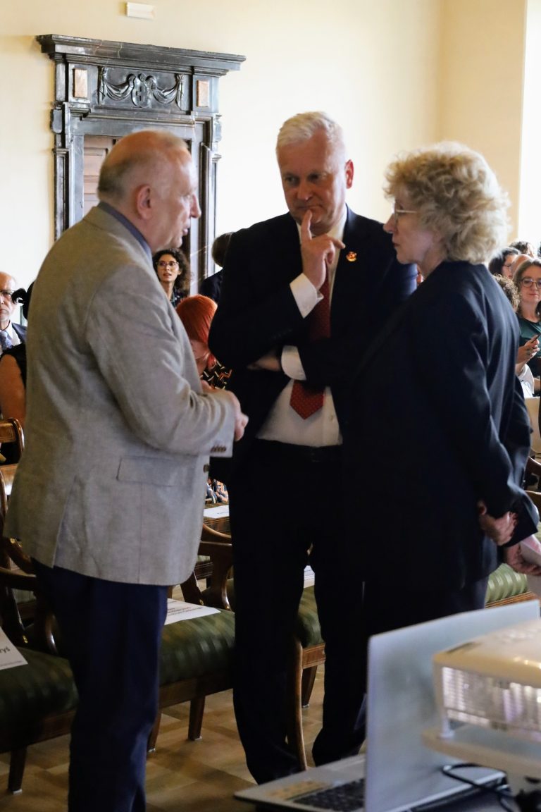 Trzy osoby prowadzą rozmowę wewnątrz budynku. Mężczyzna po lewej, ubrany w szary żakiet, gestykuluje, co sugeruje, że aktywnie wypowiada się w dyskusji. Mężczyzna w centrum, w granatowym garniturze, z zamyśloną miną i palcem na brodzie, słucha uważnie. Po prawej, kobieta z kręconymi, jasnymi włosami również patrzy na mówcę, co wskazuje na jej pełne zaangażowanie w konwersację. W tle widać fragment mebli oraz siedzące osoby.