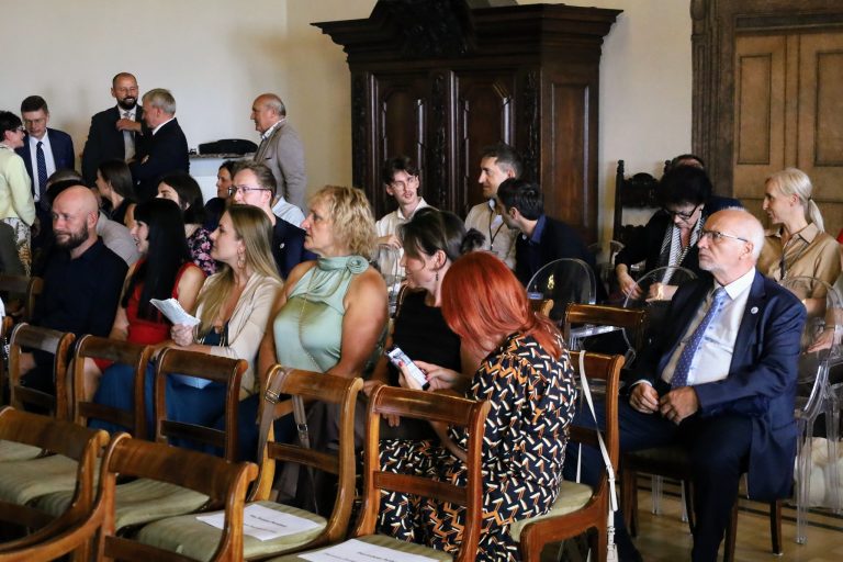 Zdjęcie przedstawia grupę osób siedzących na krzesłach w rzędach, wewnątrz eleganckiego pomieszczenia. Większość z nich siedzi z uwagą skierowaną w jednym kierunku, prawdopodobnie na scenę lub prelegenta, co sugeruje, że uczestniczą w spotkaniu lub wykładzie. Wiele osób trzyma w dłoniach programy lub notatki. W górnej części zdjęcia widać stojące osoby w trakcie swobodnej rozmowy.