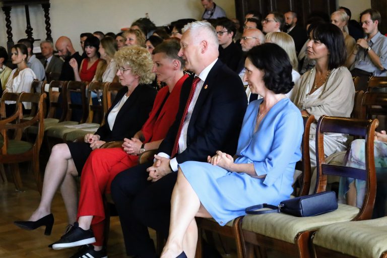 Zdjęcie przedstawia grupę osób siedzących w rzędach. Na pierwszym planie widać pięć osób, z których cztery siedzą, a jedna, mężczyzna w ciemnym garniturze i czerwonym krawacie, jest w centralnym punkcie kadru. Obok niego, po lewej stronie, siedzą dwie kobiety, a po prawej kolejna dwie. Wszyscy są elegancko ubrani, a ich uwaga skierowana jest w jednym kierunku, prawdopodobnie na scenę.