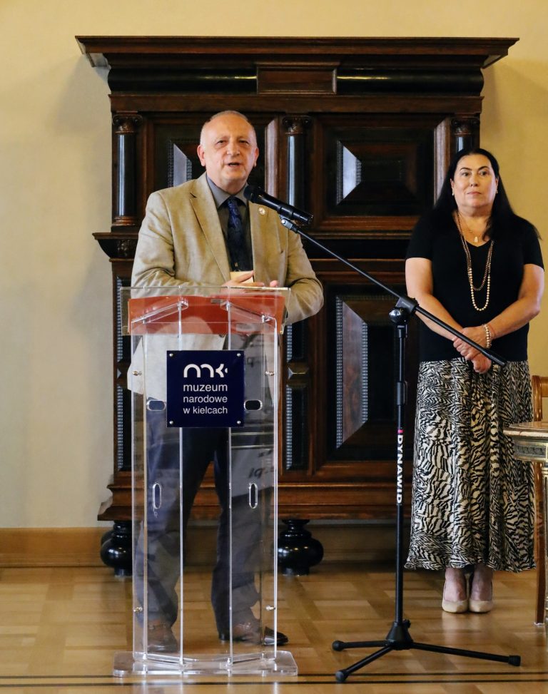 Na zdjęciu jest mężczyzna, który ma na sobie beżową marynarkę, jasną koszulę oraz krawat. Stoi za przeźroczystym, szklanym mównicą, na której widnieje logo z napisem "muzeum narodowe w kielcach". Mężczyzna przemawia do mikrofonu, a jego ręce są złożone. Obok niego, po prawej stronie zdjęcia, stoi kobieta ubrana w czarną bluzkę i spódnicę we wzór zebry. Ręce ma splecione, a na szyi ma naszyjnik z pereł. W tle, za plecami obydwu osób, widać dużą, drewnianą, zabytkową szafę, a po prawej stronie kawałek stołu.
