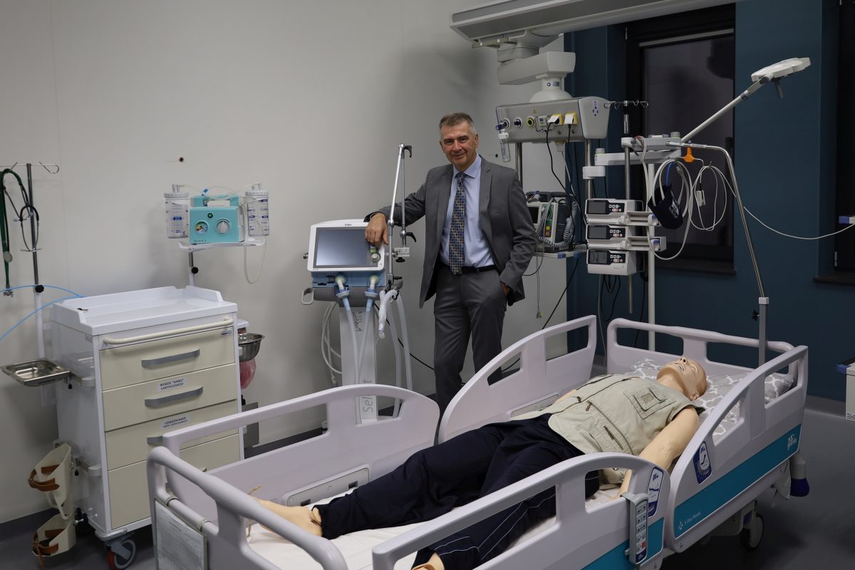 Mężczyzna w szarym garniturze stoi w sali szpitalnej lub symulacyjnej, w pobliżu sprzętu medycznego. Opiera się o dużą maszynę z ekranem dotykowym. W tle znajduje się łóżko szpitalne z manekinem. Mężczyzna patrzy wprost w obiektyw, uśmiechając się.