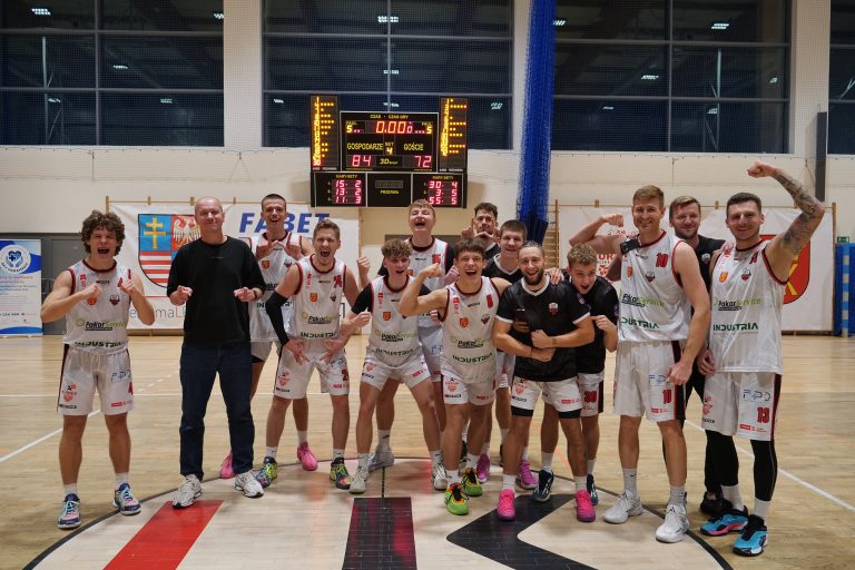 Na zdjęciu widoczna jest drużyna koszykówki świętująca zwycięstwo na boisku sportowym. Zawodnicy w białych strojach z czerwono-czarnymi elementami pozują uśmiechnięci i pełni energii. W tle widoczna jest tablica wyników – gospodarze wygrali 84:72. Wszyscy wyglądają na szczęśliwych i dumni z osiągnięcia.